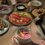 Top-down view of a wooden table spread with a variety of healthy breakfast foods, including smoothie bowls, a large platter of sliced watermelon and papaya, and pancakes.