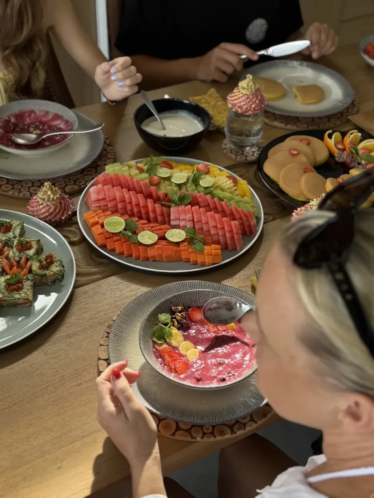 Top-down view of a wooden table spread with a variety of healthy breakfast foods, including smoothie bowls, a large platter of sliced watermelon and papaya, and pancakes.